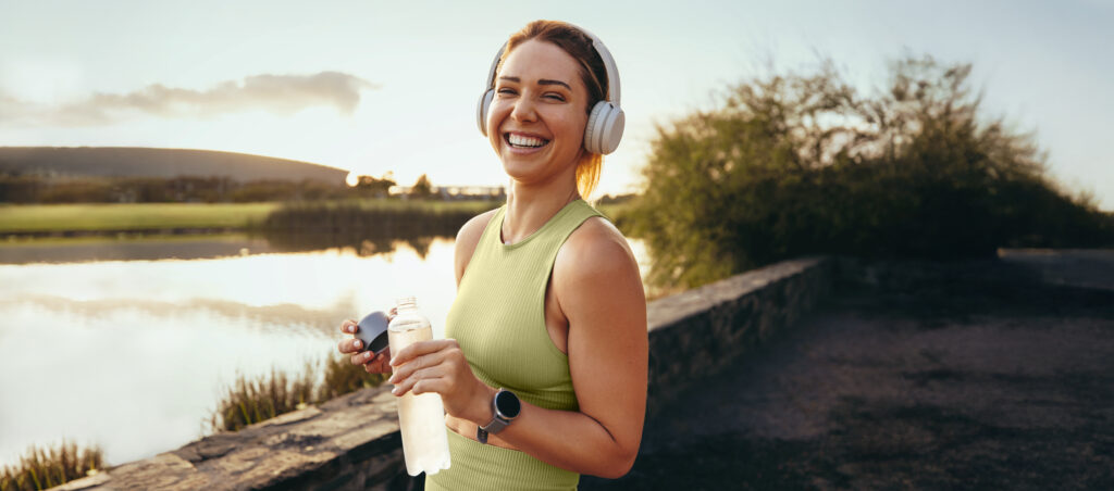 mulher sorrindo e tomando água enquanto pratica atividade física ao ar livre